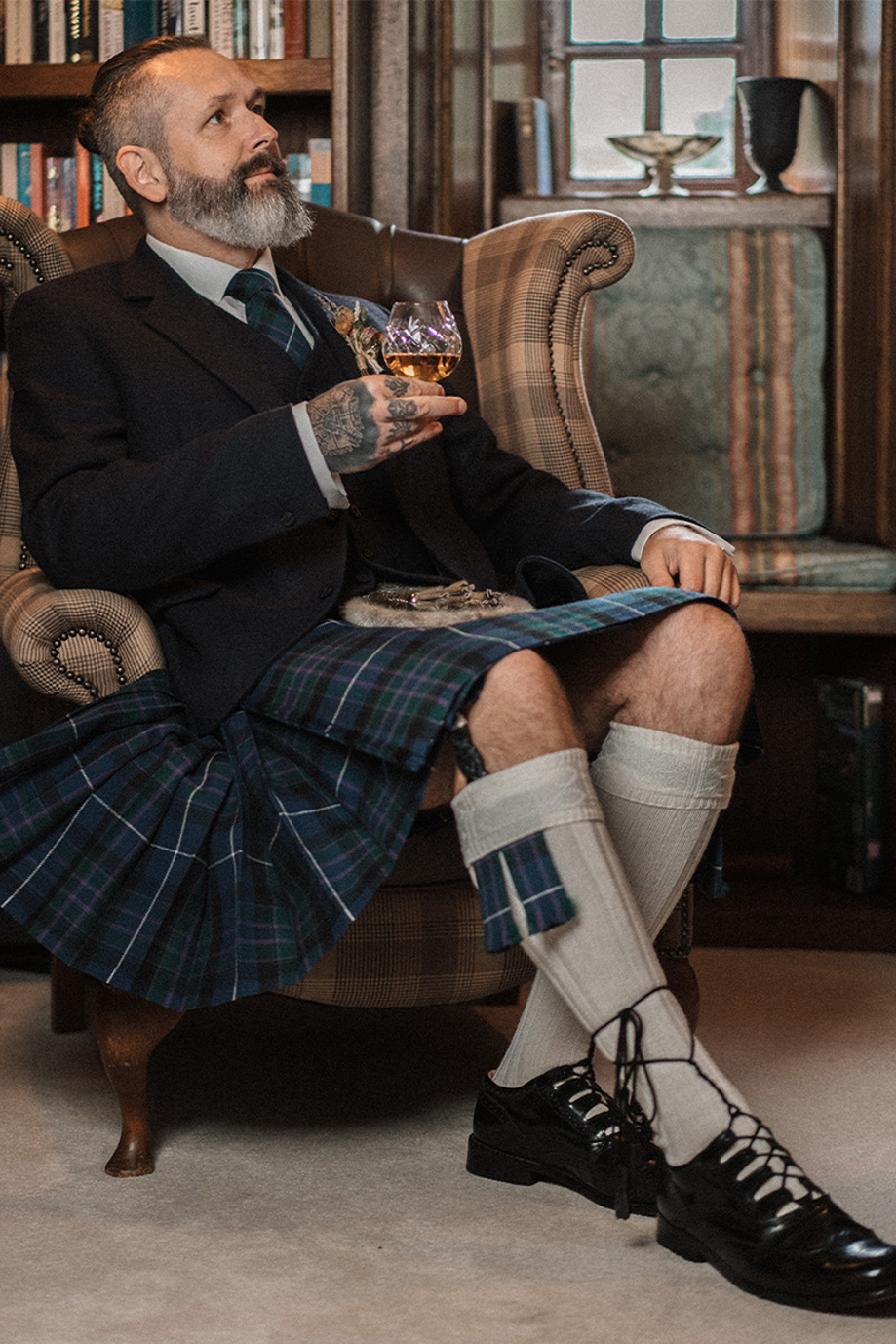 Groom in kilt sitting on tartan chair drinking whisky