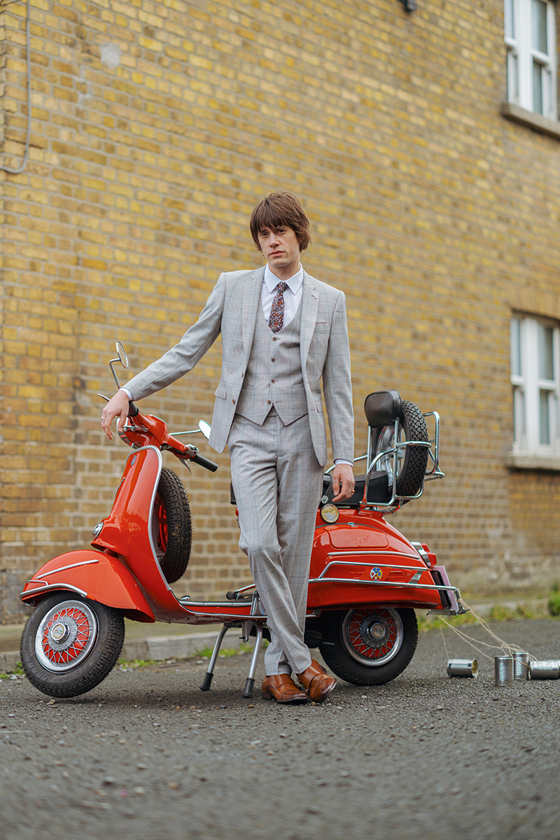 Man in grey three-piece suit leaning against red moped with cans attached to back