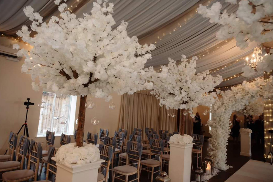 White floral trees on white pillars lining aisle, floral entrance archway with draped fabric ceiling covered in fairy lights