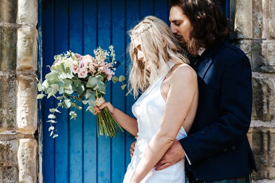 Groom stands behind bride holding bouquet with blue door in background