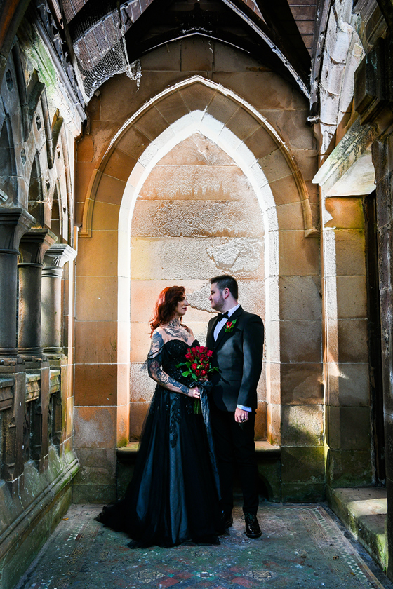 Bride and groom wearing black look at each other in front of historic arch at Geilsland Estate.