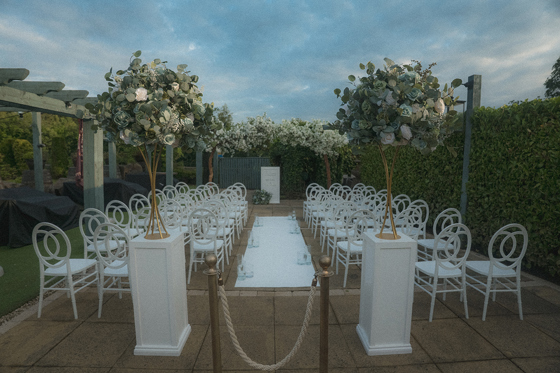 Outdoor ceremony set up with white chairs, white columns at top of aisle and floral installations.