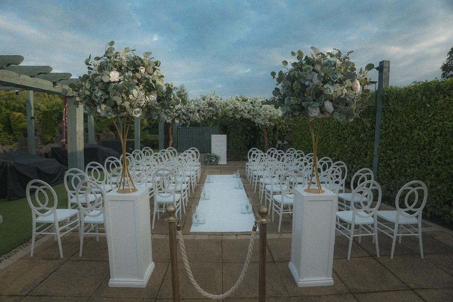 Outdoor ceremony set up with white chairs, white columns at top of aisle and floral installations.