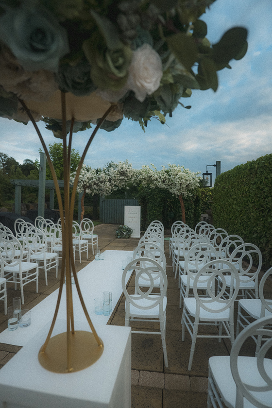 Portrait image displaying white chairs for outdoor wedding, taken from behind flowers at top of altar.