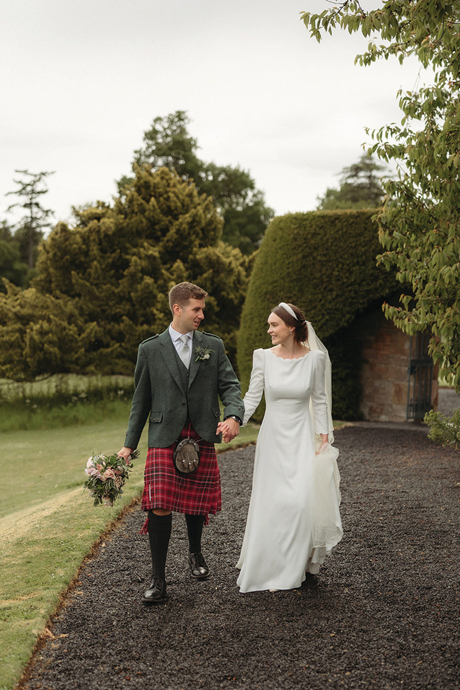 Groom holds brides bouquet as they walk along paths at Newton Don.