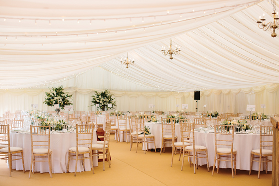 Inside a Newton Don marquee wedding meal, with round tables decorated with large floral centrepieces and gold chiavari chairs.