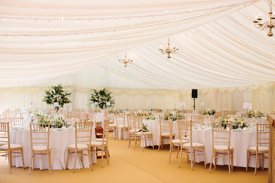 Inside a Newton Don marquee wedding meal, with round tables decorated with large floral centrepieces and gold chiavari chairs.