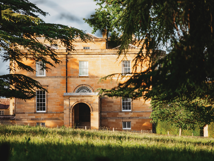 External view of Newton Don House from behind trees with tree shadows and sun shining on grass in foreground.