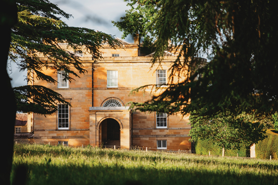External view of Newton Don House from behind trees with tree shadows and sun shining on grass in foreground.