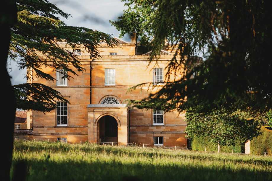 External view of Newton Don House from behind trees with tree shadows and sun shining on grass in foreground.
