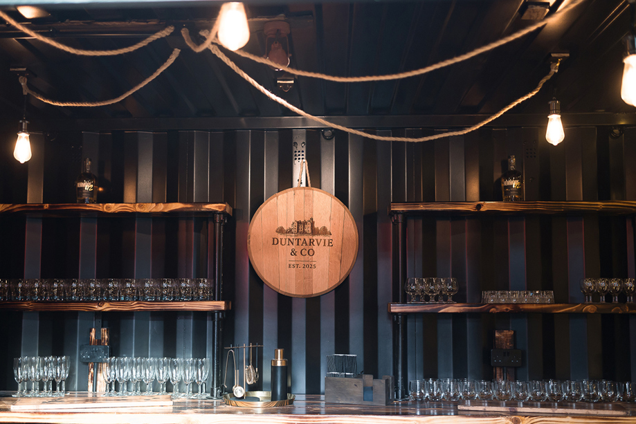 Black and wood wedding bar at The Venue @ Duntarvie Castle, featuring rustic shelving, glassware and circular Duntarvie & Co sign in modern Scottish barn venue.
