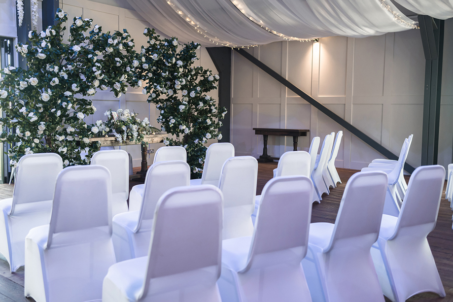 Romantic indoor ceremony space at The Venue @ Duntarvie Castle featuring floral arch backdrop, white chair covers and soft ambient lighting.