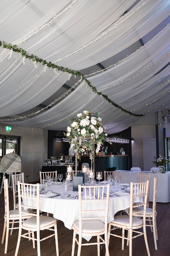 Wedding reception table styling at The Venue @ Duntarvie Castle with tall floral centrepiece, candles and cream Chiavari chairs beneath draped ceiling.