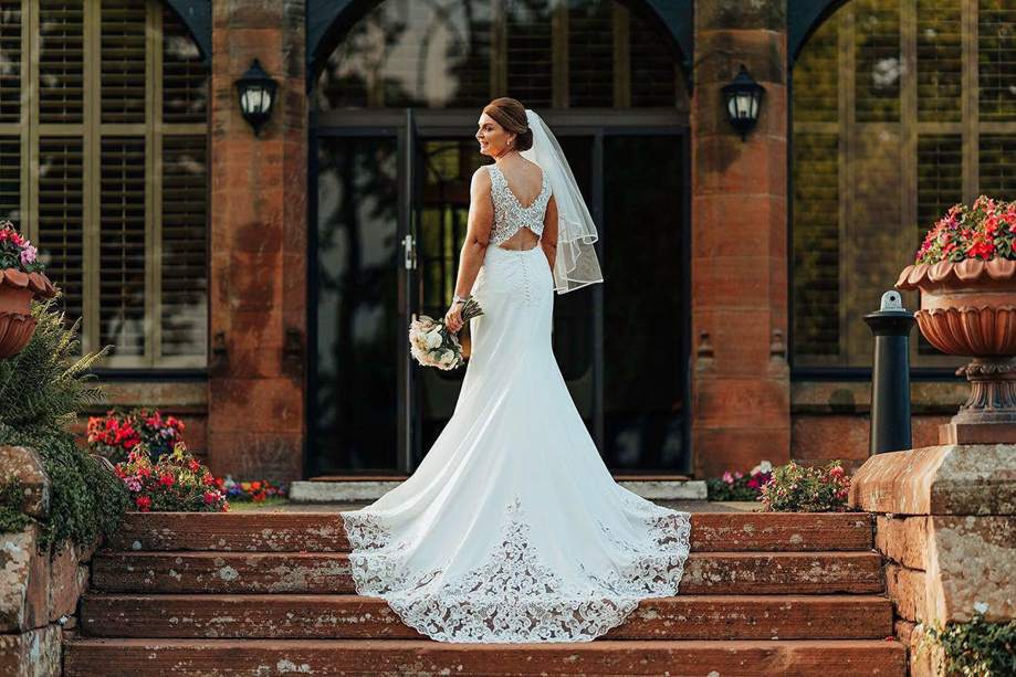 Bride standing on the steps outside Piersland House wedding venue in a lace wedding dress with long train and bouquet