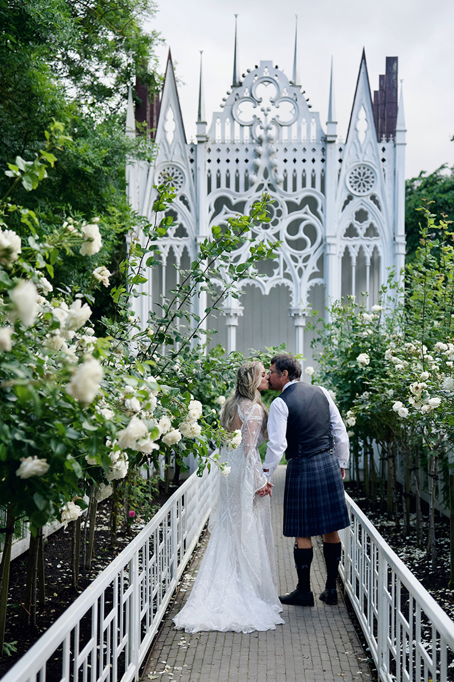 Newlywed couple kissing in the rose garden at Jupiter Artland with white sculptural installation behind them
