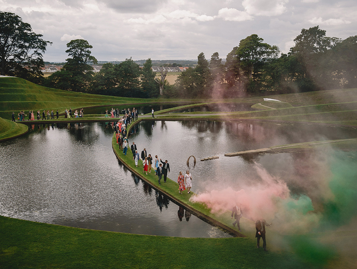 Wedding guests walking beside the sculpted ponds at Jupiter Artland with colourful smoke filling the grounds