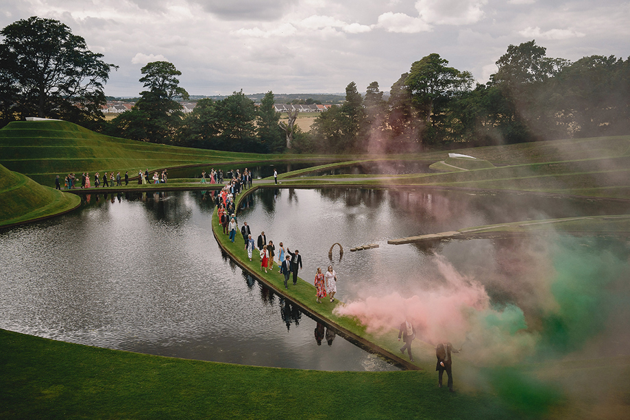 Wedding guests walking beside the sculpted ponds at Jupiter Artland with colourful smoke filling the grounds