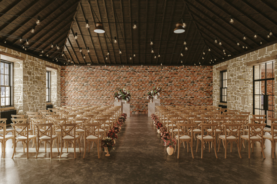 Wedding ceremony setup in the Gallery Room at The West Works Glasgow, with stone walls, brick backdrop, wooden chairs and floral aisle arrangements