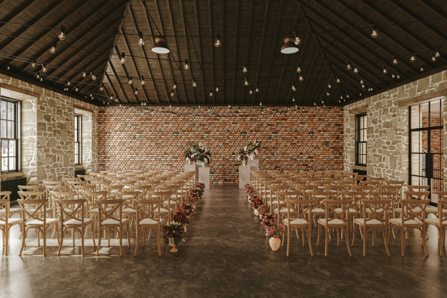 Wedding ceremony setup in the Gallery Room at The West Works Glasgow, with stone walls, brick backdrop, wooden chairs and floral aisle arrangements