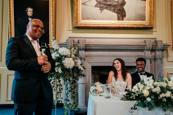Wedding guest delivers a speech with a microphone as the bride and groom laugh at the top table in Lower College Hall at the University of St Andrews.