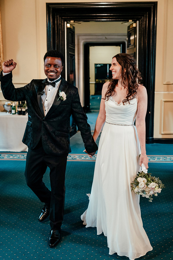Bride in a flowing white wedding dress and groom in a black tuxedo walk hand in hand into their wedding reception at the University of St Andrews.
