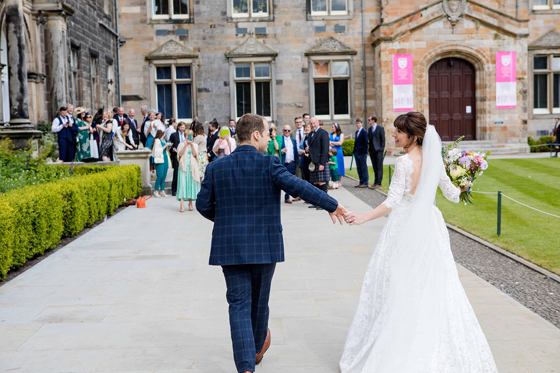 Bride in a lace wedding dress and groom in a blue checked suit walk hand in hand outside the historic University of St Andrews buildings as guests watch.