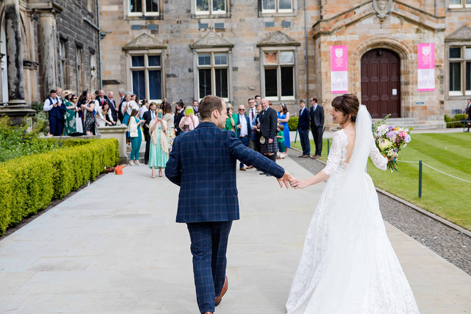 Bride in a lace wedding dress and groom in a blue checked suit walk hand in hand outside the historic University of St Andrews buildings as guests watch.