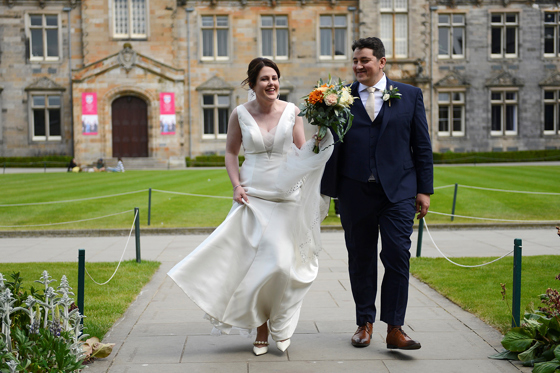 Bride carrying a colourful bouquet and groom wearing a navy three-piece suit walk together through the historic University of St Andrews courtyard.