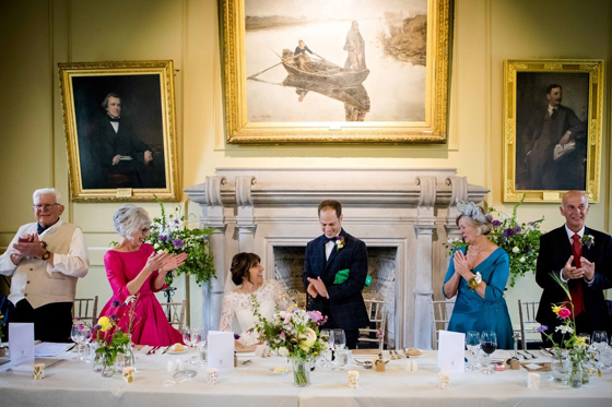 Groom stands to deliver a wedding speech as family members applaud around the top table in Lower College Hall at the University of St Andrews.