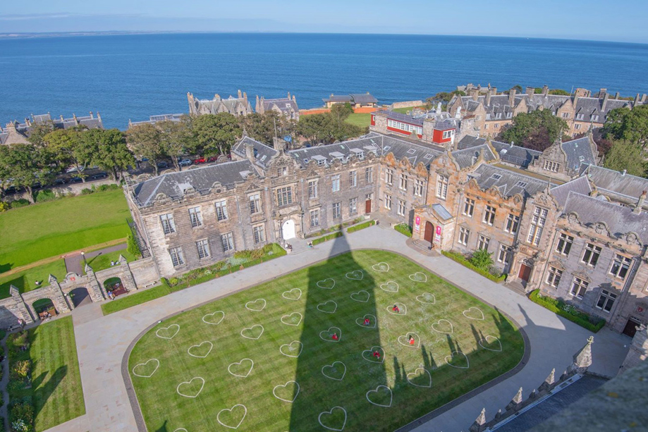 Aerial view of the University of St Andrews historic college buildings and courtyard lawn decorated with heart shapes, with the North Sea beyond.