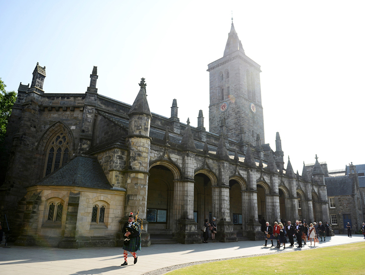 Traditional Scottish bagpiper leads a wedding party past the historic St Salvator's Chapel at the University of St Andrews.