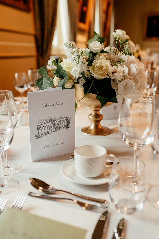 Close-up of a wedding breakfast table setting with white floral centrepiece, gold vase, personalised stationery and glassware at the University of St Andrews.