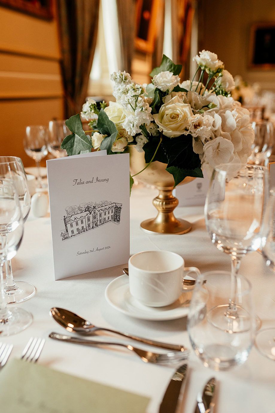Close-up of a wedding breakfast table setting with white floral centrepiece, gold vase, personalised stationery and glassware at the University of St Andrews.