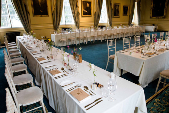 Long wedding breakfast tables dressed with white linen, delicate flowers and white Chiavari chairs in a historic reception hall at the University of St Andrews.
