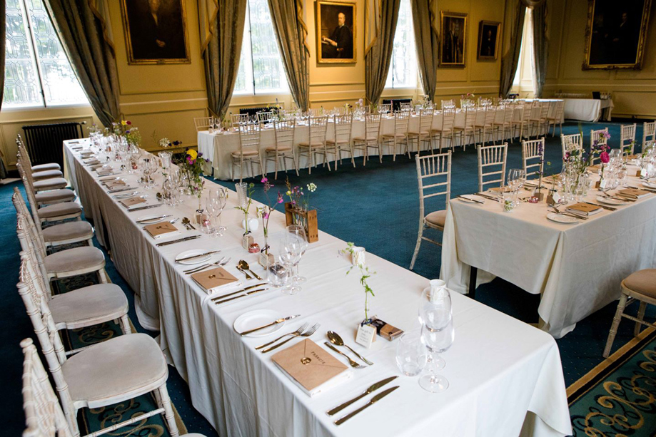 Long wedding breakfast tables dressed with white linen, delicate flowers and white Chiavari chairs in a historic reception hall at the University of St Andrews.