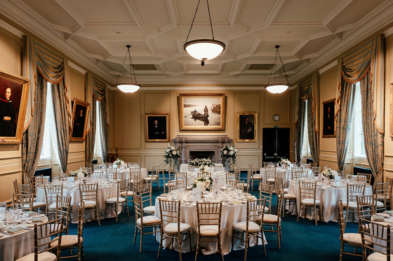 Lower College Hall at the University of St Andrews set for a wedding breakfast with round tables, gold Chiavari chairs, white flowers and historic portraits.