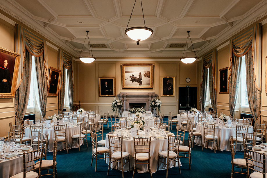 Lower College Hall at the University of St Andrews set for a wedding breakfast with round tables, gold Chiavari chairs, white flowers and historic portraits.