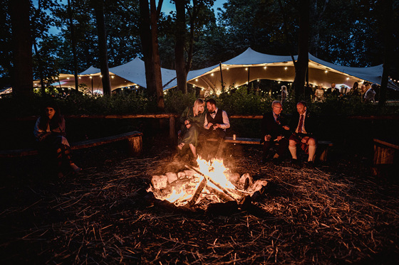 Bride and groom relax by the bonfire with tent covered in fairy lights in background