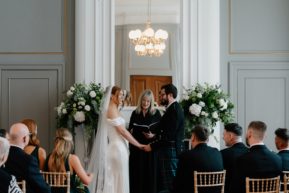 Bride and groom during their wedding ceremony at The Edinburgh Grand, framed by white and green floral arrangements.