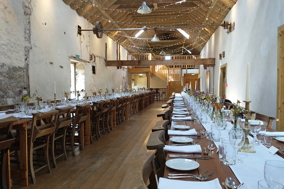 Long trestle tables set for a wedding meal inside Cormiston Farm barn with exposed beams