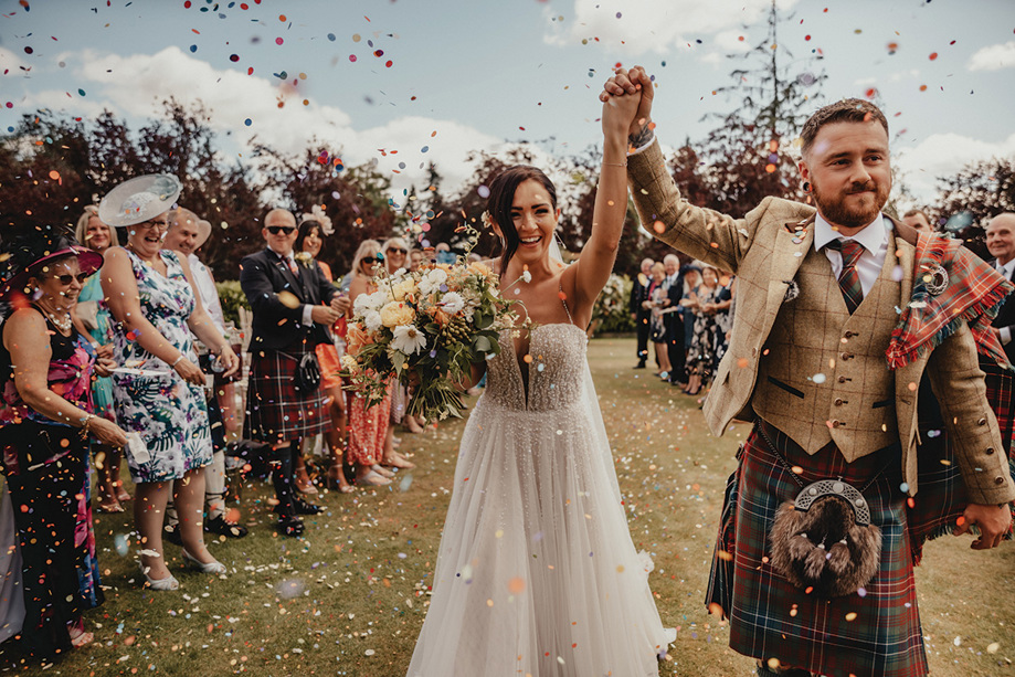 Bride and groom hold their hands up in air as they walk through confetti after getting married