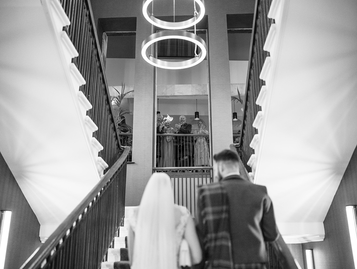 Black and white image of a bride and groom walking upstairs at The Clayton Hotel in Glasgow city centre, while guests look on and clap at the top.