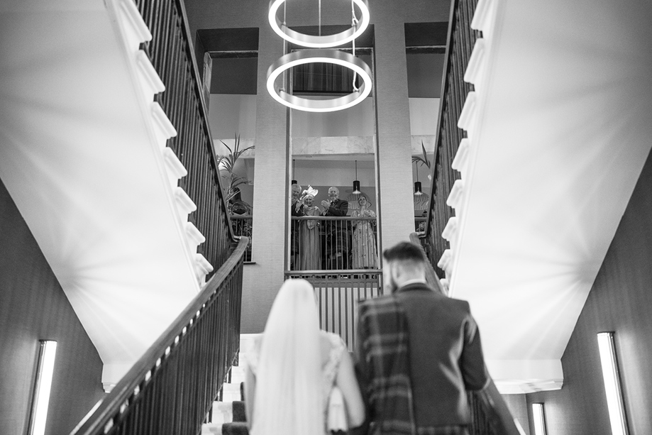 Black and white image of a bride and groom walking upstairs at The Clayton Hotel in Glasgow city centre, while guests look on and clap at the top.