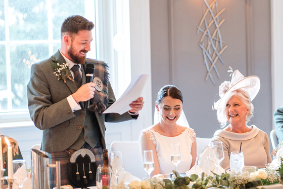 Groom makes speech at top table while bride and mother of the bride laugh at their wedding breakfast at The Clayton Hotel in Glasgow city centre