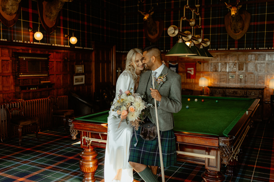 Bride and groom leaning on a pool table inside Dornoch Station Hotel billiards room, groom wearing tartan kilt and bride holding bouquet
