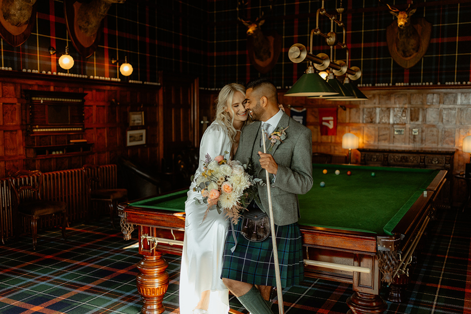 Bride and groom leaning on a pool table inside Dornoch Station Hotel billiards room, groom wearing tartan kilt and bride holding bouquet