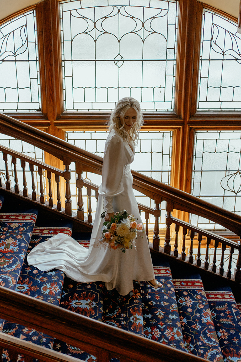 Bride walking down ornate staircase at Dornoch Station Hotel holding bouquet, with stained glass windows and patterned carpet backdrop