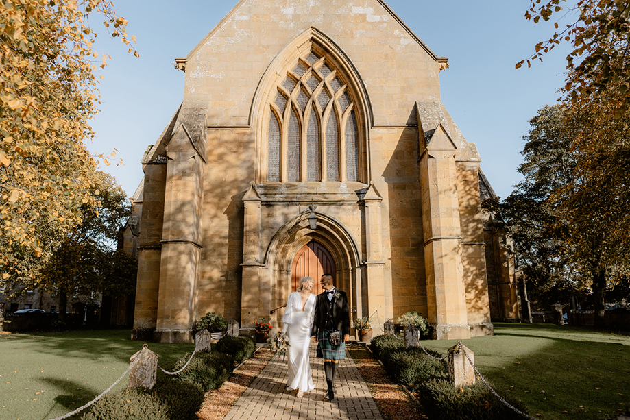 Newly married couple walking hand in hand outside historic cathedral near Dornoch Station Hotel wedding venue