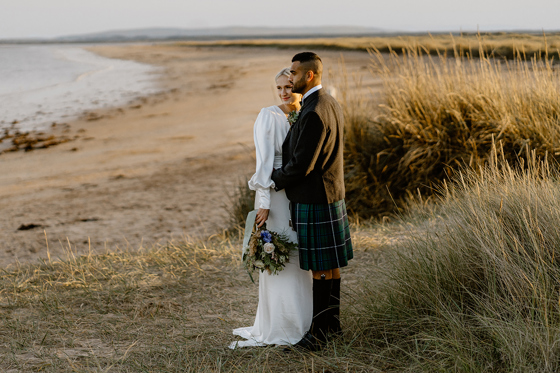 Bride and groom standing together on sandy beach near Dornoch, overlooking coastal grass dunes during Scottish Highlands wedding photos