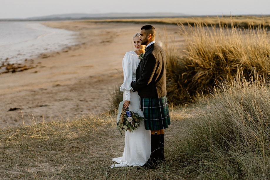 Bride and groom standing together on sandy beach near Dornoch, overlooking coastal grass dunes during Scottish Highlands wedding photos